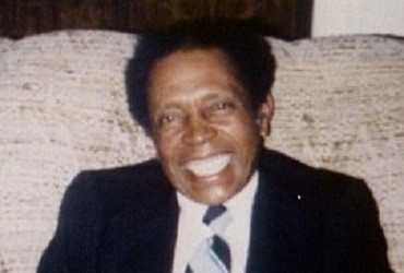 Rogest Cain smiling at the camera, sitting on a couch in a dark blue suit