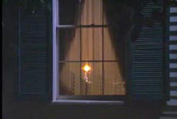 A candle lit in a window with sheer curtains and blue shutters.