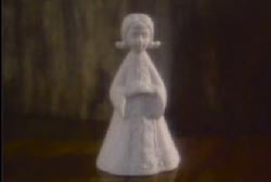 A small white porclein bell in the shape of child angel sitting on a wood table.