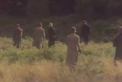 roup of men in hats and trenchcoats searching through a field. 
