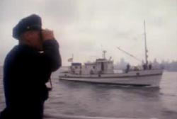 A police officer searches the water through binoculars, there is a coast guard boat in the background.