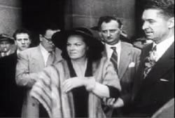 Virginia Hill wearing a fur scarf, hat and gloves. She is flanked by several men in suits.