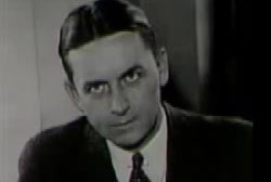 Eliot Ness sitting on a chair staring at the camera in a black suit.