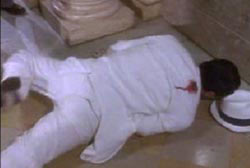 A man in a white suit is laying face down on the ground, he is bleeding from a gun shot wound in his right shoulder.