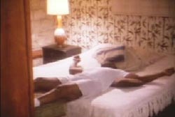 Sonny Liston is sprawled on a bed, he is wearing a white t-shirt, boxers and socks. There is a lamp on the bedside table.