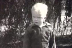 A small boy with blonde hair is standing wearing a jumpsuit.