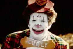 A clown in white makeup, a red flannel suit and a bowtow that says 'Curly'