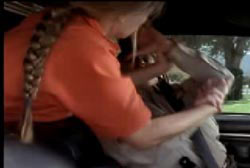 A woman with a braid and an orange shirt is reacing over the back seat to attack an officer.