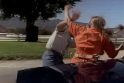 A woman in an orange jumpsuit is attacking another person next to a car.