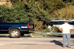 A man in a white tshirt and jeans stands in front of a blue pick up truck towing a boat.