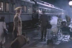 Two uniformed soldiers are on a train platform. One is walking away while the other is watching him.