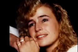 A young woman is posing, reasting her heads on her hands, her blonde hair is curly and she is wearing a white shirt.