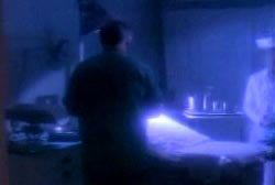 An autopsy room with a man in shadow standing over a corpse on a gurney.