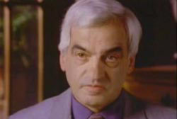 An elderly caucasian man with grey hair wearing a purple suit.