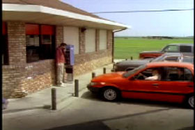 A red sedan parked next to a brick and mortar store. There is a man on a red shirt on a payphone.