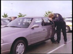 A police officer peering into a grey sedan abandoned in a parking lot.