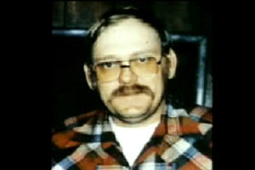 A middle aged man with a mustache and orange tinted glasses, Lee 'Dub' Wackerhagen, wearing a plaid shirt.