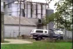 A blue and white pick up truck parked infront of farm silos.