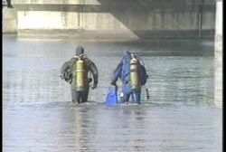 Two men in scuba gear wade into the water under a bridge