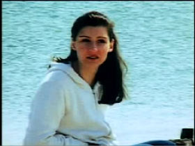 Claudia Kirschhoch wearing a oversized white sweater, she is sitting on a beach with the water behind her.