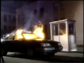 A sedan is on fire in front of security booth on a parking lot.