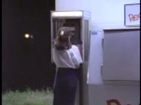 A woman in a white sweater and jeans standing at a payphone.