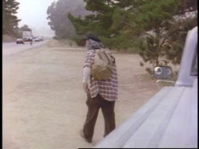 A man in a plaid shirt, brown pants and a cap walk away from a car parked on the side of the road.