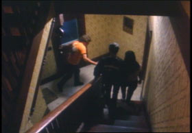 Two people walking down the stairs towards the entrance of a building as a man in orange shirt enters.