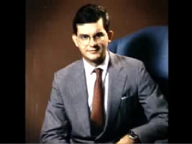 A middle aged caucasian man wearing wire rim glasses and a suit sitting in an armchair, John Cheek.