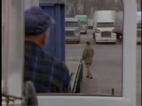A man in a purple plaid shirt watches as a man in a green jacket walks through a parking lot at a truck stop.