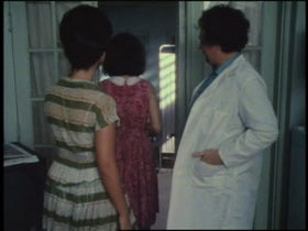 Two women walk past a doctor in a lab coat into an examine room.