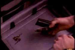 A hand reaches into the truck of the car and picks up a wallet, there are keys laying next to the wallet.