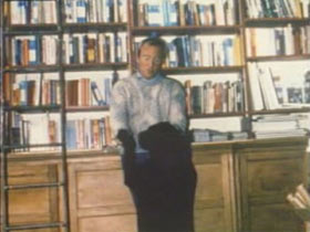 A man dancing with a black dog in front of a large bookshelf.