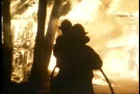 Two firefighters are aiming a firehose at a house engulfed in flame.