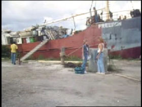 Three people standing on a dock infront of a freighter. The word 'Freedon' is painted on the bow of the ship.