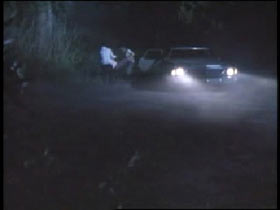 A dark sedan with it's headlights on a wooded road at night. Two men are lifting something out of the trunk.