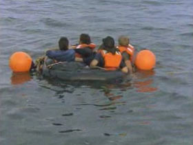 Four men packed into a small life raft, there are two large orange bouys attached to the raft. 