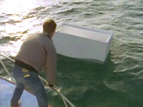 A man is standing on a boat, leaning over the railing trying to reach a large white box that is floating.