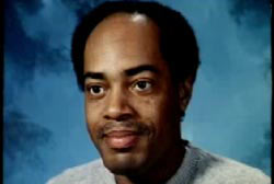 An African American, Oliver Munson. He has a receding hairline and is posing in front of a blue bakcdrop.