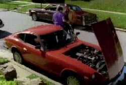 Two men stand next to a red compact car, the hood is open and the engine is visible.