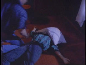 A person is laying on the floor with a white pillow over their face as another person kneels next to them.