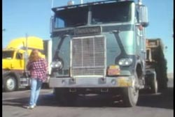 A woman in a purple plaid shirt walking past the front of a semitruck.