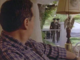 A man in a plaid shirt is sitting in his living room. He is looking through the window to see two military men approaching the front door.