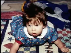 A baby, Sabrina Aisenberg, in a blue jumpsuit playing on a blanket.
