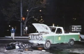 A green and white pick up truck in the middle of the street, the hood is up and the front of the truck is damaged.