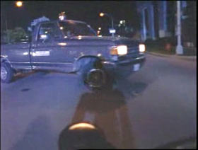 A person on a motorcycle is stopped when a dark pick up truck pulls infront of it.
