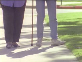 Two people walking through a park, we can see them for the waist down and one is using a cane.