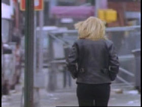 A woman with blonde hair and a leather jacket walking away down a street.