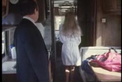 A woman in a robe walks down the hall of a mobile home as a man in suit watches her.