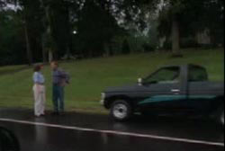 Two people standing in front of a pick up truck that is parked on the shoulder of a street.
